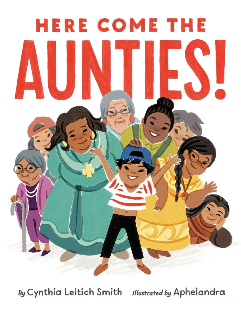 The book cover for Here Come the Aunties! By Cynthia Leitich Smith, illustrated by Aphelandra. A young Muscogee child stands with his hands up and a big smile in front of a group of diverse women of all ages who are smiling over his shoulder.